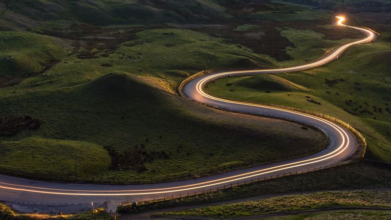 Das Foto zeigt eine sich durch grüne Hügel schlängelnde Landstraße, entlang der eine Lichtspur von Scheinwerfern zu sehen ist.
