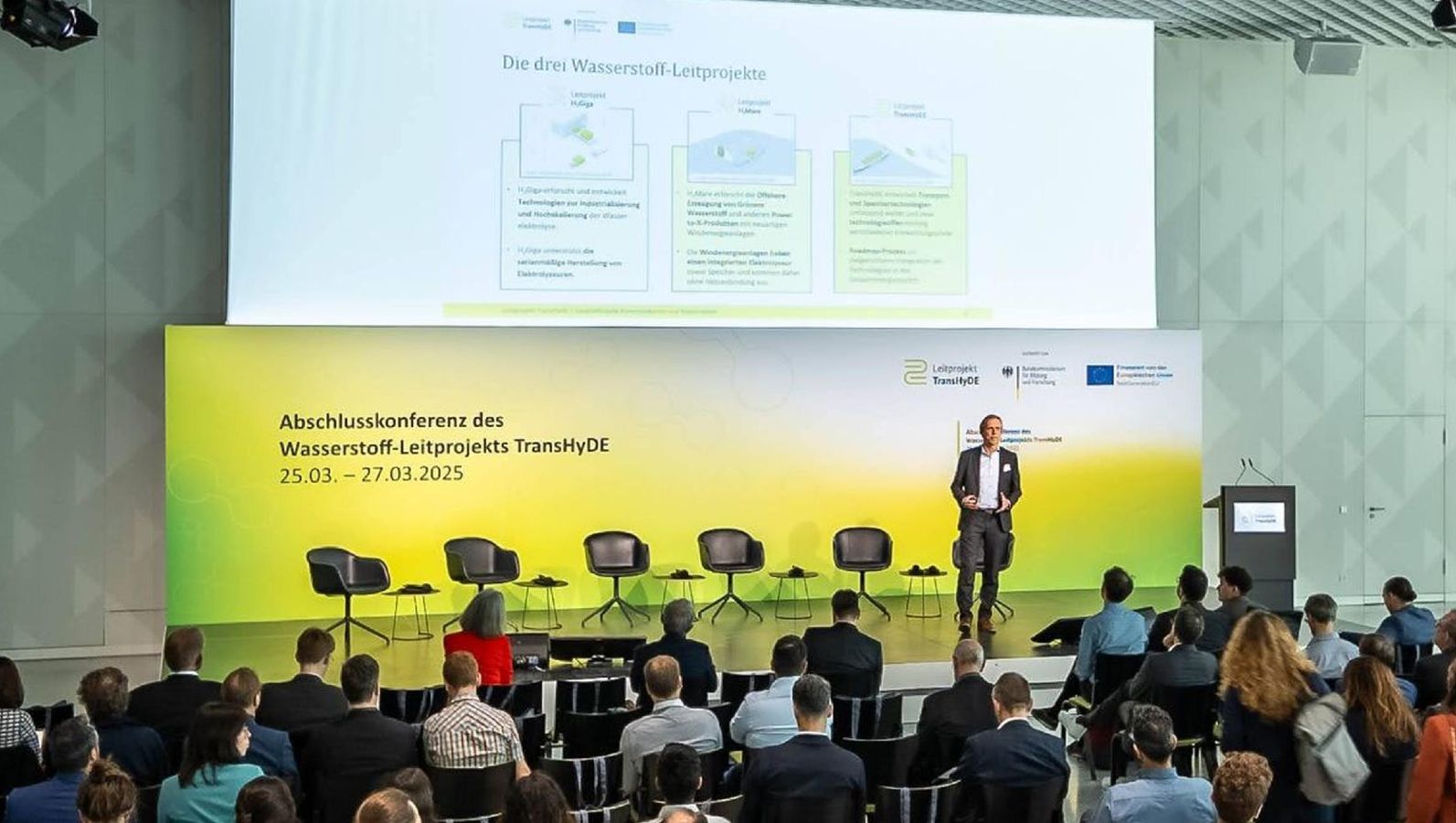 Das Bild zeigt die Bühne der Abschlusskonferenz. Im Hintergrund Leinwände mit Infos zu den Leitprojekten, im Vordergrund Teilnehmende der Konferenz.