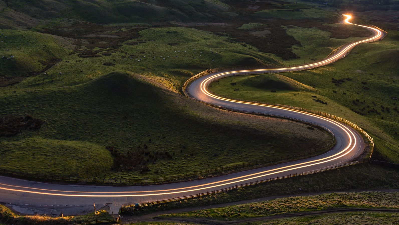Das Foto zeigt eine sich durch grüne Hügel schlängelnde Landstraße, entlang der eine Lichtspur von Scheinwerfern zu sehen ist.