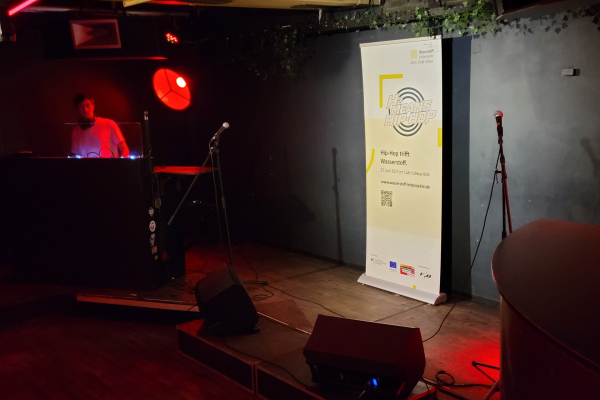 Das Bild zeigt die Bühne im Kölner Club Subway mit einem Rollup des Hip-Hop-Events. Daneben ist der DJ zu sehen.